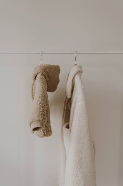 Sonbahar kıyafetleri beyaz duvardan sarkıyor. Sıcak bebek ve yetişkin ceketleri. Kıyafetleri tarafsız renklerde olan estetik bir gardırop.