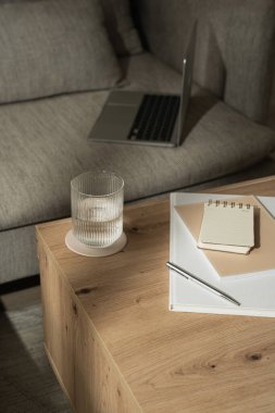 Evde çalış. Dizüstü bilgisayarlı estetik ofis çalışma alanı, bir bardak su, defter, belgeler, güneş ışığı gölgeli ahşap masa üzerinde fotokopi alanı olan boş kağıt kağıtlar.
