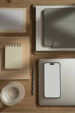 Boş ekranlı cep telefonu, dizüstü bilgisayarı, tableti, defteri, bir bardak suyu olan düz konutlu ofis çalışma alanı. Sabah işi, iş konsepti. Masada güneş ışığı gölgeleri var. Düz konum, üst görünüm, kopyalama alanı