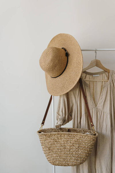 Women's fashion clothes and accessories. Stylish female straw handbag, straw hat, beige linen dress on hanger over white background. Minimalist fashion blog