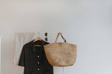 Beyaz duvara asılmış zarif kadın kıyafetleri. Siyah gömlek, pantolon, güneş gözlüğü, rattan çanta. Yazlık kıyafetlerle moda kompozisyonu