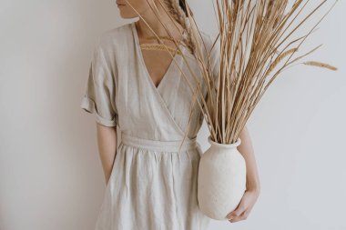 Estetik kum tenceresi ve kadın ellerinde kurutulmuş pampa otu. Beyaz duvarın üzerinde, bej bej bir elbise içinde genç, güzel bir kadın var. Boho, bohem moda tarzı.