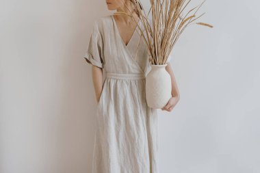 Kumlu bej elbiseli kadın, beyaz duvarında kurutulmuş pampa otları olan toprak takımlarını tutuyor. Estetik boho, bohem stili moda konsepti