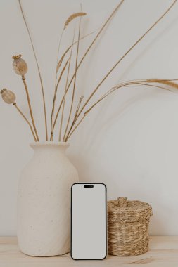 Boş ekranlı cep telefonu, rattan tabutu, kuru otlu kil çömlek ve beyaz duvarda haşhaş sapı. Sosyal medya için estetik nötr bej şablonu, model için kopyalama alanı olan mobil web sitesi