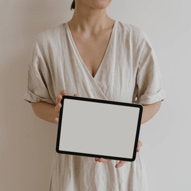 Tarafsız bej keten elbiseli güzel bir kadın beyaz duvarda boş ekranlı tablet taşıyor. Sosyal medya için estetik şablon, model için fotokopi alanı olan mobil web sitesi