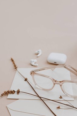 Gözlükler, kablosuz kulaklıklar, kurutulmuş çimenler, kağıt çarşaflar, zarf. Nötr bronz bej masa üzerindeki minimalist çalışma masası kırtasiyesi. Estetik yaratıcılık kavramı
