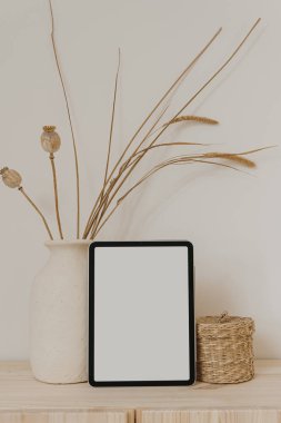 Boş ekranlı tablet tableti, kuru otlu kil tabut ve beyaz duvarda haşhaş sapı. Sosyal medya için estetik nötr bej şablonu, model için kopyalama alanı olan mobil web sitesi