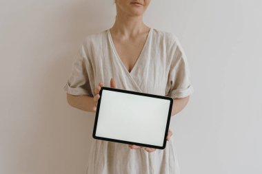 Tablet kadınların elinde. Beyaz duvarın üzerinde, bej bej bir elbise içinde genç, güzel bir kadın var. Boho, bohem tarzı. Boş boş kopya alanı boş ped ekran ile estetik maket
