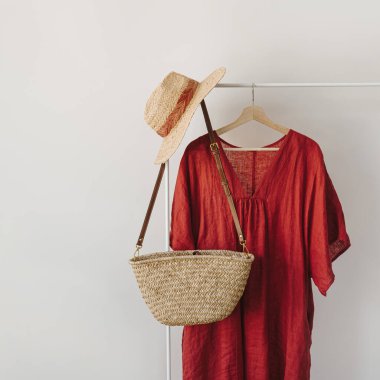 Kadın kıyafetleri ve aksesuarları. Zarif kadın hasır çanta, hasır şapka, beyaz arka planda asılı kırmızı elbise. Minimalist moda günlüğü