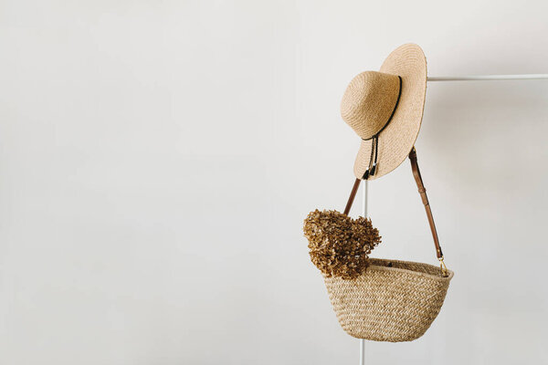 Women's fashion accessories. Stylish female straw handbag, straw hat and dried flower on hanger over white background. Minimalist fashion blog
