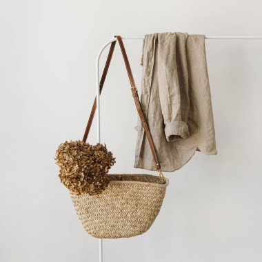 Zarif moda aksesuarı. Kurumuş ortanca çiçekli saman çanta, askıda bej keten gömlek. Moda günlüğü, kopyalama alanı olan sosyal medya şablonu