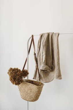 Hasır şapka, saman çanta, bej keten gömlek, kurutulmuş ortanca çiçeği beyaz duvarın üzerinde asılı.