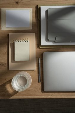 Evde çalış. Laptop bilgisayarı, tablet tableti, bir bardak su, defter, belgeler, ahşap masa üzerinde güneş ışığı gölgeleri olan boş kağıt kağıtlı estetik ofis çalışma alanı.