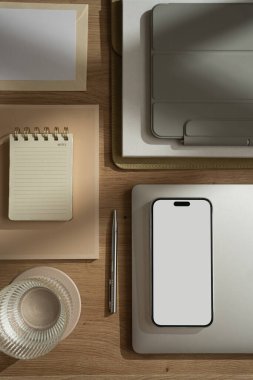 Boş ekranlı cep telefonu, dizüstü bilgisayarı, bir bardak su, tablet, güneş ışığı gölgeli defter olan ofis çalışma alanı. Estetik lüks bohem iş hayatı konsepti. Düz yatış, üst görünüm