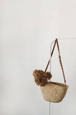 Saman el çantası, kurutulmuş ortanca çiçeği beyaz duvarın üzerinde elbise askılığı.