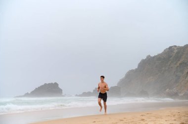 Atletik yapılı genç bir adam sahilde yalınayak koşar. Praia da Ursa. Portekiz, 2024. Yüksek kalite fotoğraf