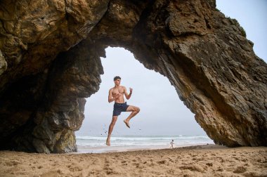 Atletik yapılı genç bir adam havaya sıçrıyor ve kaslarını geriyor. Praia da Ursa. Portekiz, 2024. Yüksek kalite fotoğraf