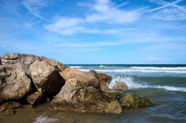 Güneşli bir günde deniz kıyısında kayalık bir sahil. Deniz dalgaları kayalık kıyıya çarpıyor.