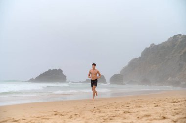 Atletik yapılı genç bir adam sahilde yalınayak koşar. Praia da Ursa. Portekiz, 2024. Yüksek kalite fotoğraf