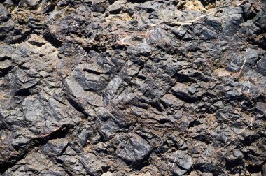 Karmaşık desenler ve dokular, güneş ışığıyla aydınlatılan kayalık bir oluşumun karanlık yüzeyinde görülebilir. Engebeli arazi, bu bölgede bulunan jeolojik çeşitliliği gösteriyor..