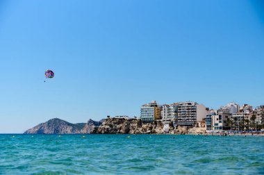 Canlı bir paraşüt, modern binalar ve kıyı bölgesindeki berrak mavi gökyüzü ile kaplı güzel kıyı şeridini gözler önüne sererek köpüklü suların üzerinde yüzer. Benidorm, Alicante, İspanya 10.01.2023