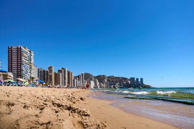 İnsanlar Benidorm, İspanya 'da bir plajda sıcak güneşin tadını çıkarıyorlar. Yüksek binalar parlak mavi gökyüzüne ve nazik dalgalara karşı çarpıcı bir zemin oluşturur. Benidorm, Alicante, İspanya 10.01.2023