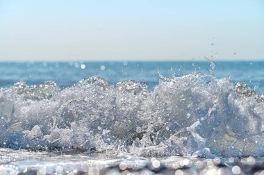 Su, parlak güneş ışığıyla aydınlatılan kumlu sahile dalgalar akarken enerjik bir şekilde sıçrıyor. Ufuk, sakin bir mavi tonla uzanıyor ve huzurlu bir atmosfer yaratıyor..