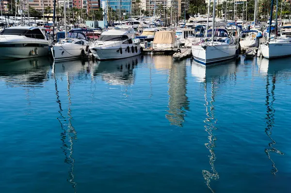 Birçok yat ve tekne bir marinada demirli. Su berrak ve sakin, parlak mavi gökyüzünün altındaki damarları ve yakınlardaki binaları yansıtıyor. Alicante, İspanya 10.01.2023