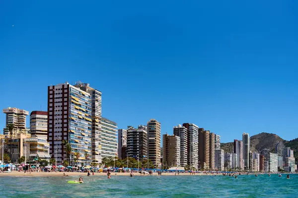 Altın kumsal, modern yüksek binalar kıyı şeridi boyunca uzanan güneş banyosu ve yüzücülerle doludur. Açık mavi gökyüzü bu popüler sahil beldesinin canlı atmosferini artırır. Benidorm, Alicante, İspanya 10.01.2023