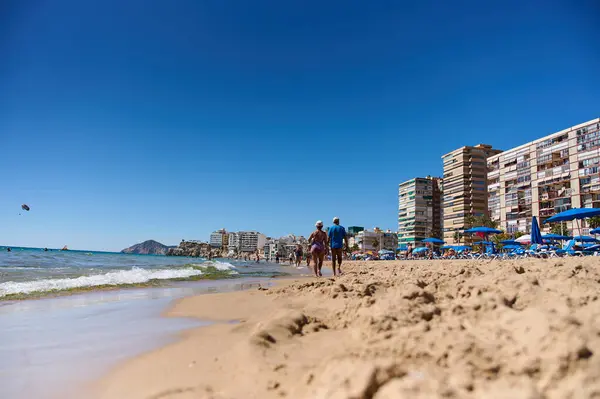 İki kişi kumlu bir plajda el ele yürürken, dalgalar yavaşça kıyıya vururken ve binalar yaklaşırken güneşli havanın keyfini çıkarırken. Benidorm, Alicante, İspanya 10.01.2023