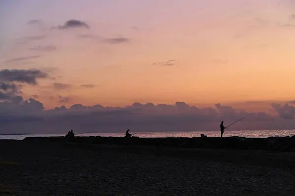 Güneş ufkun ötesinde batarken, balıkçılar canlı renklere karşı siluetlerine bürünmüş, sabırla avlanmalarını bekliyorlar. Huzurlu atmosfer barışçıl yansımayı davet ediyor..