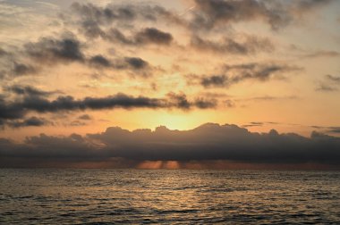 Güneş ufkun altına batarken, altın renkler gökyüzüne yayıldı, turuncu ve pembenin derin gölgeleriyle bulutları aydınlattı. Okyanus sükunetteki güzelliği yansıtıyor, huzuru davet eden anları..