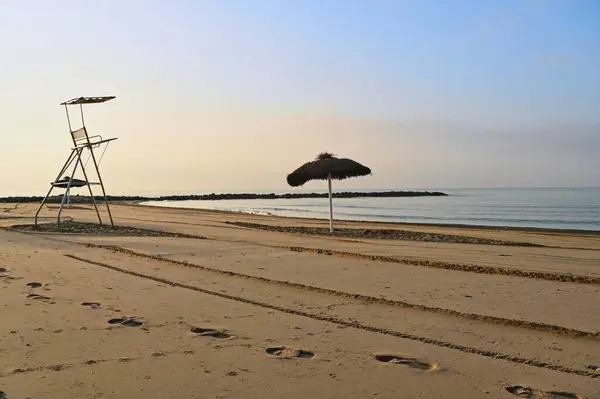 Ayak izleri altın kumu aşıp bir şemsiyenin altındaki cankurtaran sandalyesine götürüyor. Sakin dalgalar kıyıya vurur, yumuşak sabah ışığında yıkanır, huzurlu bir atmosfer yaratır..