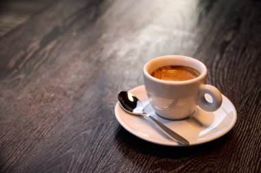 Bir fincan dumanı tüten kahve, gümüş bir kaşıkla birlikte küçük beyaz bir tabakta duruyor, şık bir kafe ortamında sıcaklık ve sükuneti çağrıştırıyor, yansıtmak için mükemmel..