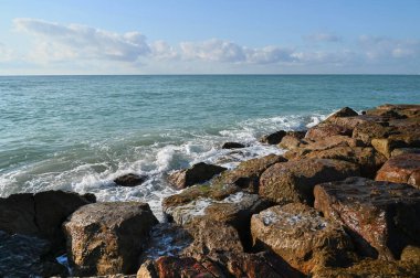 Güneş okyanusun sakin sularını aydınlatırken küçük dalgalar kıyı boyunca uzanan yıpranmış taşlarda ritmik olarak tur atar. Yumuşak bulutlar yukarıdaki geniş gökyüzünde.