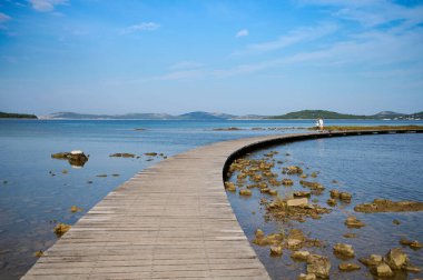 Sakin suların üzerinden ahşap bir patika esiyor, keşifleri davet ediyor. Yakınlardaki kayalar yumuşak akıntıya karşı barışçıl bir kontrastlık yaratırken, tepeler ufuk çizgisini siluete çeviriyor..
