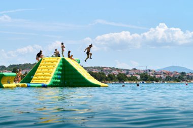 Heyecanlı bireyler canlı, yeşil şişme bir kayaktan kristal berrak sulara atlıyor, güneşli ve güneşli bir öğleden sonra sahilde eğlenceli bir günün tadını çıkarıyorlar. 07.06. 2022 Hırvatistan, Sibenik
