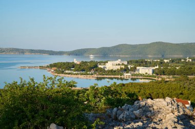 Güneş ışığı, sakin suları ve kıyı şeridinin verimli yeşilliğini nazikçe aydınlatır. Yumuşak dalgalar kıyıya vurur ve bu huzurlu ortamda barışçıl bir ortam yaratır. 07.06. 2022 Hırvatistan, Sibenik