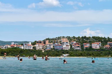 Sıcak güneş ışığı, insanların köpüklü suda yürüdüğü, güldüğü ve oynadığı sahili yıkıyor. Kıyı binaları, canlı atmosferi zenginleştiren pitoresk bir zemin oluşturur.. 