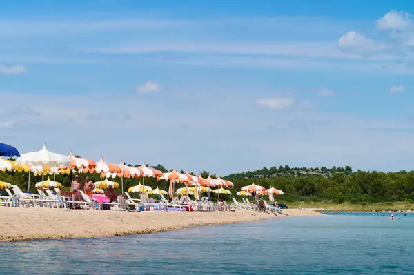 İnsanlar neşeli şemsiyelerin altında turkuaz suyun altında güneşli bir plaj gününün tadını çıkarıyorlar. Canlı atmosfer, sıcak yaz atmosferini ve kaygısız boş zamanları çağrıştırıyor. 07.06. 2022 Hırvatistan, Sibenik