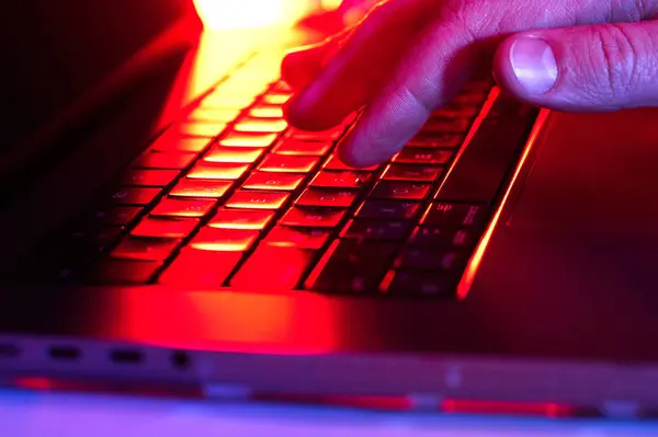 Canlı kırmızı ve turuncu ışıklarla aydınlatılan bir dizüstü bilgisayar klavyesinin üzerinde asılı duran bir el, gece yarısı verimliliğini ima ediyor..
