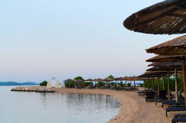 Yumuşak kum, saman şemsiyeleri altında sakin sularla buluşur. Alacakaranlık sakin bir sahile çökerken..