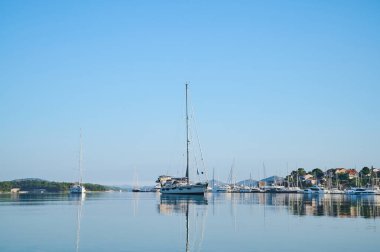 Çok sayıda yelkenli sakin bir körfezde dinleniyor. Gökyüzü açık ve su yansıyor..