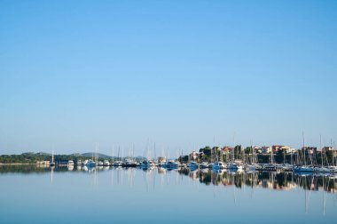 Serene marina sahnesinde açık mavi gökyüzünün altında durgun suya yansıyan tekne ve binalar var..