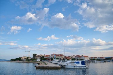 Sakin limanda çeşitli tekneler mavi gökyüzünün altında tüylü bulutlarla dolu tuhaf bir sahil köyünün yakınına yanaştı..