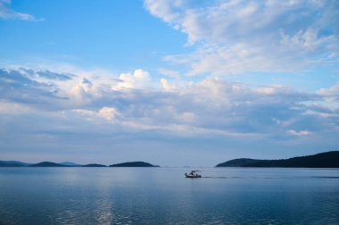 Küçük bir tekne sakin sularda, adalar ve akşamları yumuşak mavi gökyüzü ile çevrili bir şekilde hareket eder..