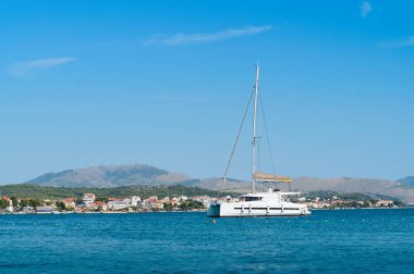 Beyaz bir yelkenli tekne sakin sularda dinlenir. Arka planda güzel bir kıyı şeridi ve dağlar vardır..