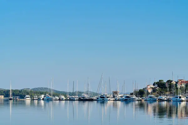 Yatlar, yeşil tepelerle çevrili, şafak vakti berrak mavi gökyüzünü yansıtan sakin bir marinaya demirlenmiştir..