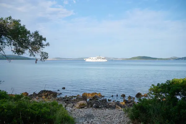 Büyük bir yat sakin sularda süzülüyor, manzaralı sahil tepeleri ve yeşilliklerle çevrili..