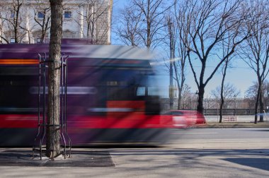 Parlak mavi bir gökyüzünün altında ağaçlarla kaplı canlı bir caddede renkli bir tramvay hızla ilerliyor..